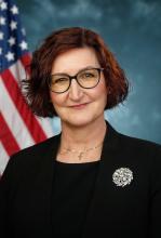 Official Photo of AnnMarie Highsmith. Woman with short hair, wearing dark glasses posing in front of an American flag wearing a black suit