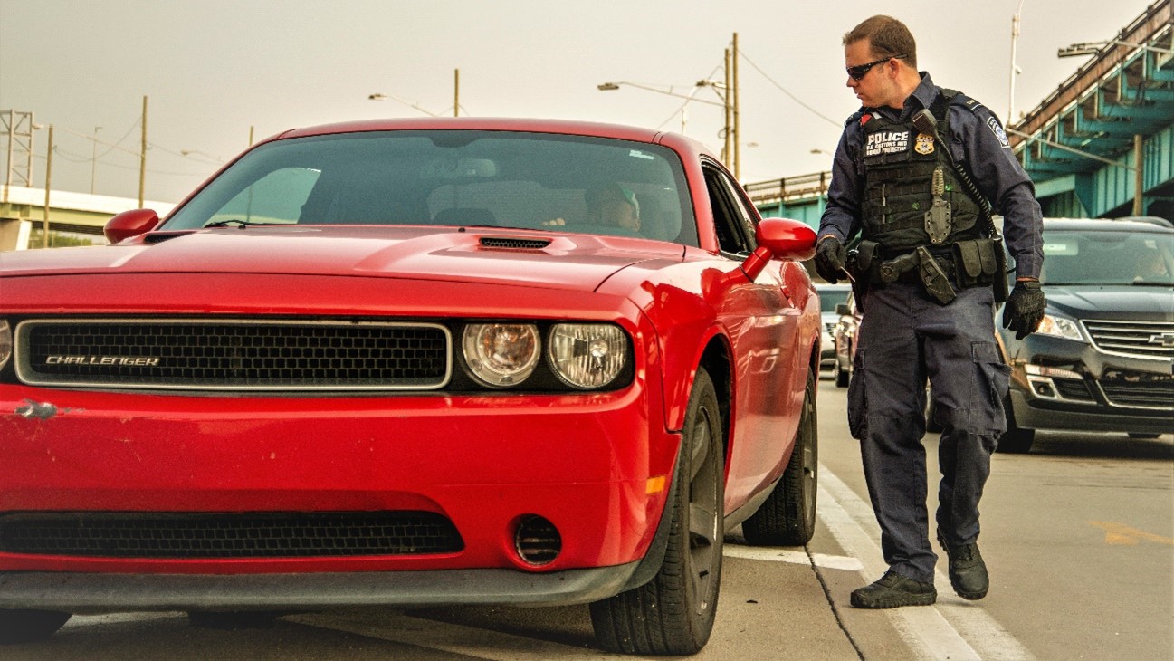 A CBP officer stands next to a vehicle and driver entering the U.S. at a port of entry.