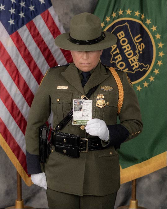 USBP agent holding a card displaying a fallen agent standing in front of the U.S. flag and U.S. Border Patrol flag
