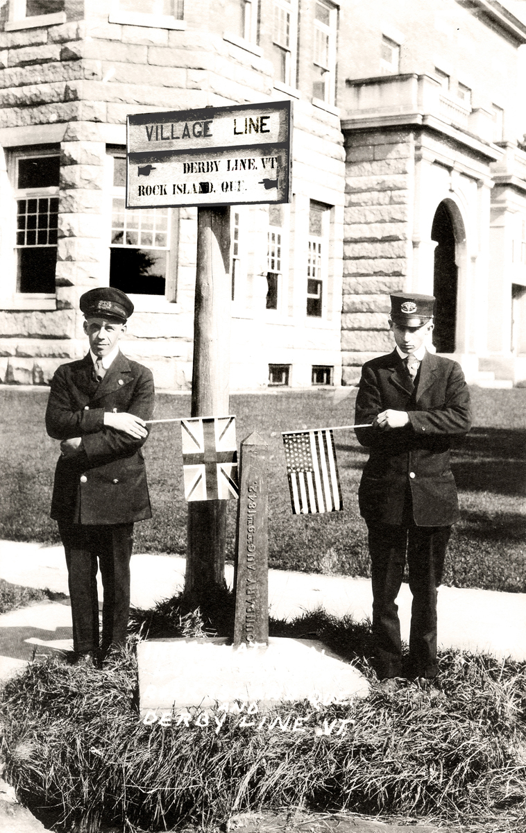 The establishment of a port of entry in Derby Line, Vt., is the result of the relocation of the Customs officer from the town of Derby northward to the international border - quite similar to the movement of the port of entry from the town of Franklin to Morses Line. In this postcard photo, Customs Inspector Ray Phelps (right) stands on the U.S. side of the boundary marker and his Canadian counterpart stands on the Rock Island, P.Q. side. In the background is the famous Haskell Free Library and Opera House that was specifically built straddling the line in order to promote cross-border friendship and community between the Vermonters and Quebecers.