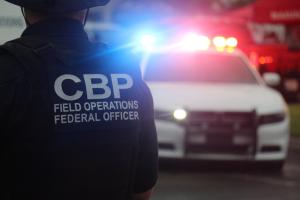 A uniformed CBP officer flanks a marked CBP unit with flashing lights.