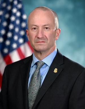 Troy A. Miller, CBP Acting Commissioner smiling in front of a blue background with a flag to the left and right of the acting.