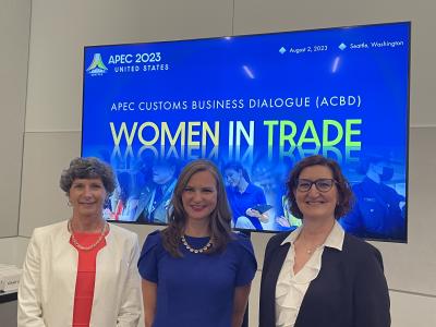 three women in suits at a trade event, posing in front of the 2023 APEC Customs sign