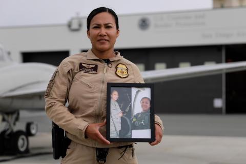 A CBP Air and Marine Operations Agent holds a picture of herself in her military uniform, while standing in front of a aircraft and a hanger.