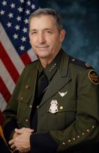 Benjamine “Carry” Huffman, CBP Acting Deputy Commissioner smiling in front of a blue background with a flag to the left and right of the acting.